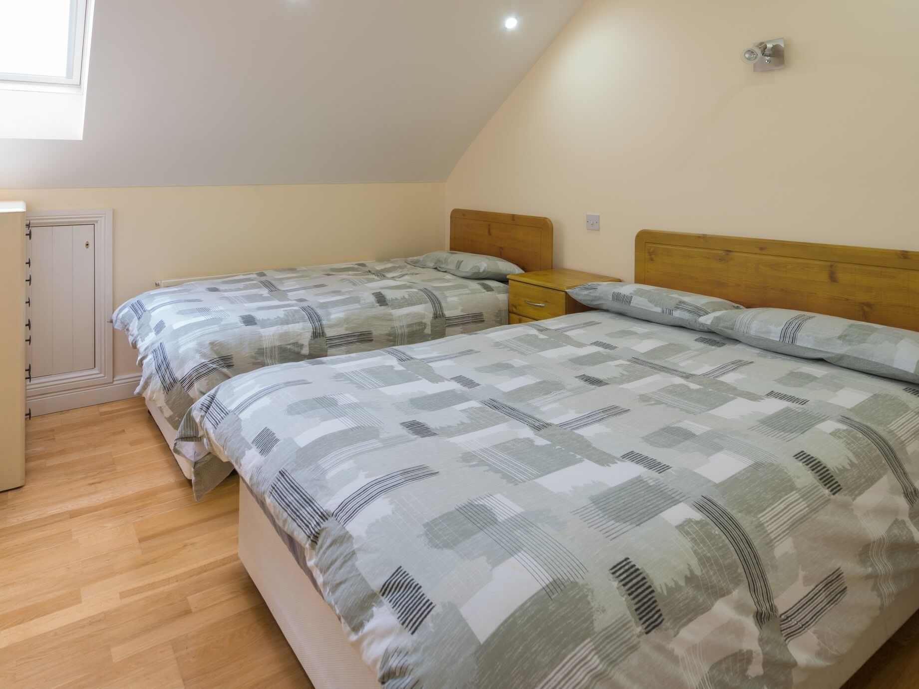 A bedroom with two beds, wooden headboards, a small bedside table, and a window.