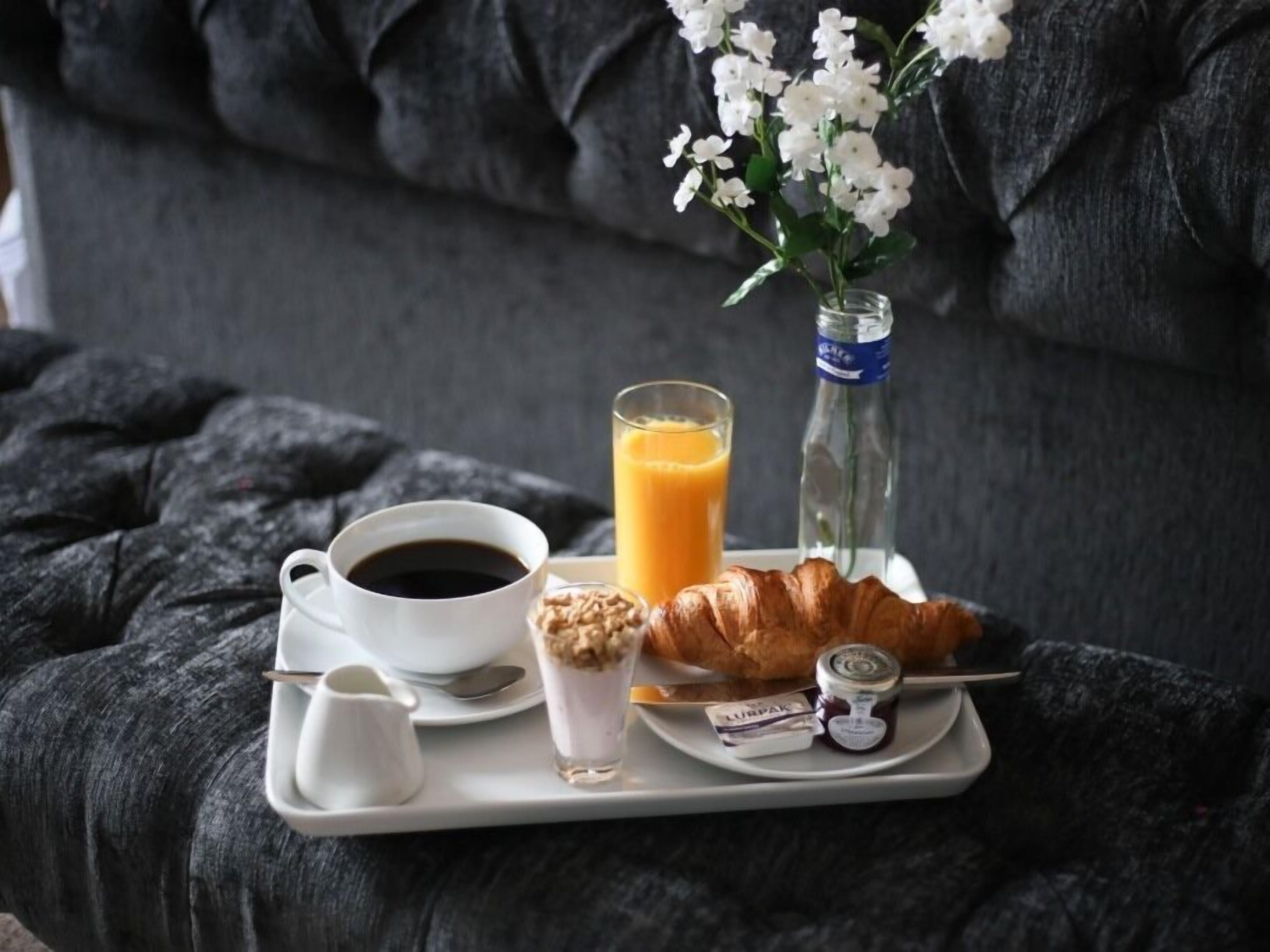 A breakfast tray with a croissant, a glass of orange juice, a cup of coffee, and a small jar of jam on a bed with a dark gray blanket.
