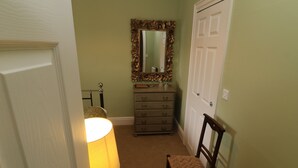 Un couloir étroit avec une chaise en bois, une commode à tiroirs, un miroir doté d’un cadre orné et une porte.