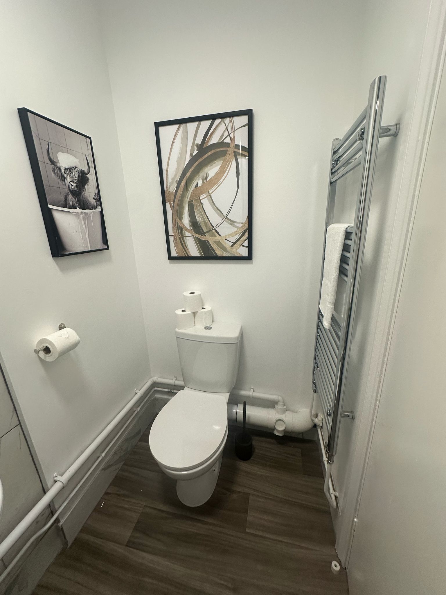A small, modern bathroom with a toilet, a hand dryer, and a radiator. There are two framed pictures on the wall.