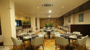 A spacious dining area with multiple tables and chairs, a patterned carpet, and a large plant in the center. - Vybrant Hotels (Vijayawada)