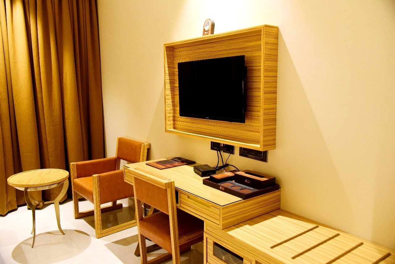 Habitación de hotel con un escritorio de madera, un televisor montado en la pared, una silla, una mesita y una ventana con cortinas.