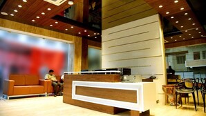 A modern reception area with a wooden reception desk, a seating area with a sofa, and a large window with a view of the cityscape. - Vybrant Hotels (Vijayawada)