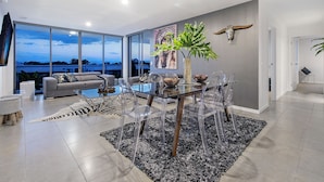 Una sala moderna con una mesa de comedor de vidrio, sillas transparentes, un sofá gris y un amplio ventanal con vistas al cielo y al paisaje.