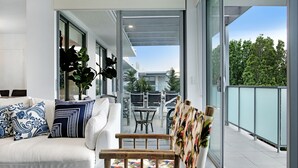Una sala moderna con un sofá blanco, cojines estampados y una silla de madera con asiento floral. Unos puertas de cristal dan a un patio exterior con una mesa y sillas.