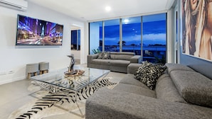 Un moderno salón con un televisor de pantalla plana grande, una mesa de centro de cristal, un sofá gris y un ventanal con vistas a la ciudad.