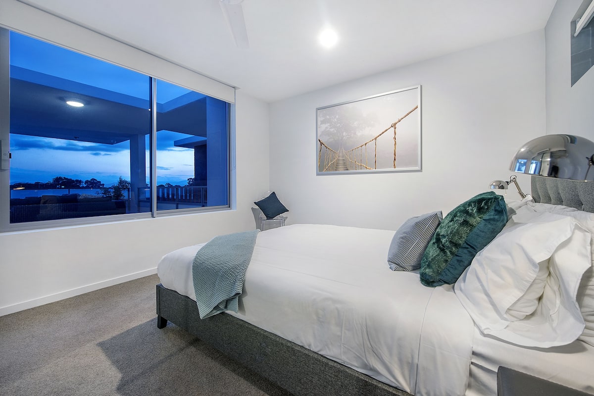 Un dormitorio moderno con un ventanal grande, una cama con ropa de cama blanca, vistas al exterior y un cuadro en la pared.