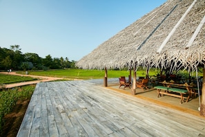 A wooden deck with picnic tables and benches, a thatched-roof pavilion, and a view of a green field. - Amuna Ayurveda and Wellness Retreat (Sigiriya)