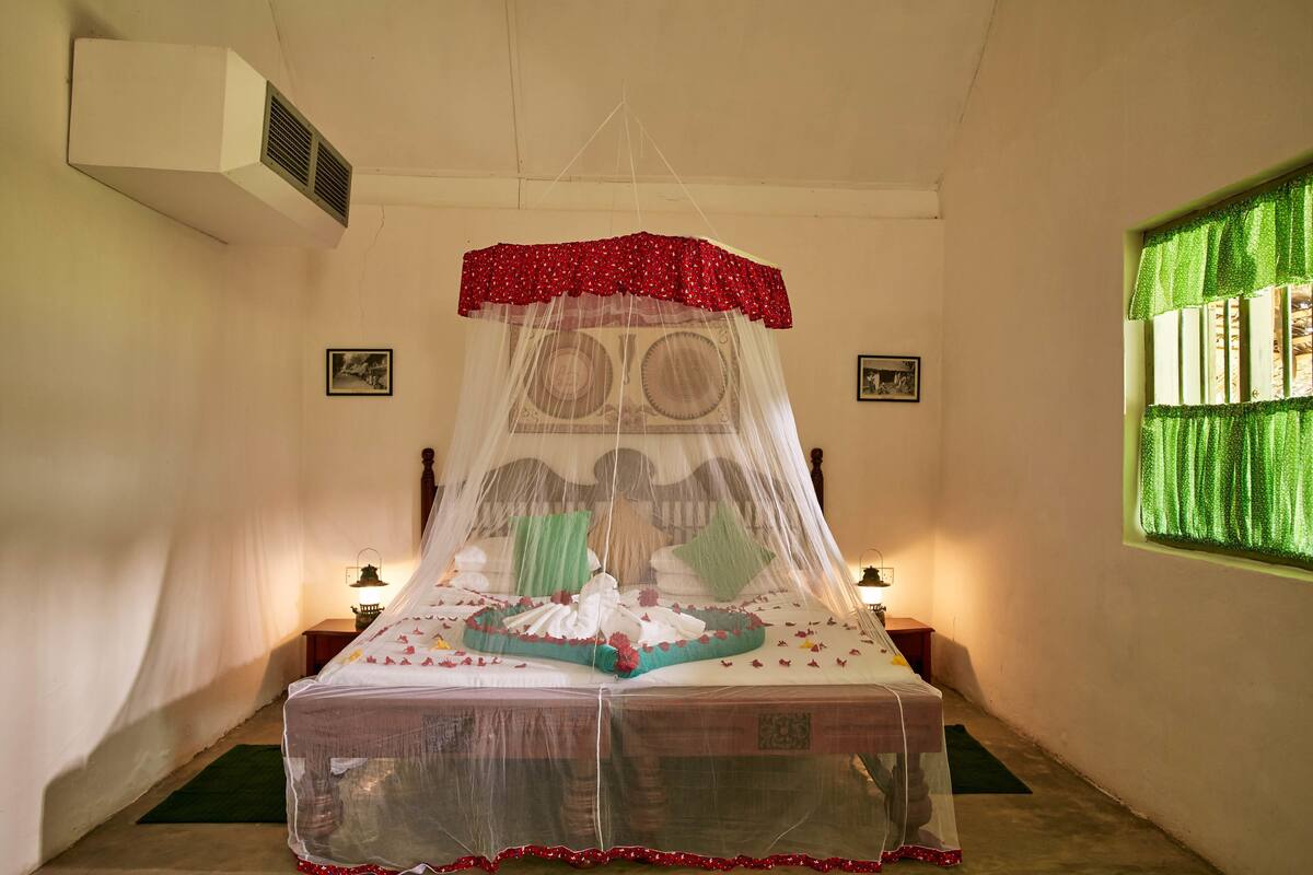 Een bed met hemelbed, een nachtkastje, een wandairconditioner en een raam met groene gordijnen.