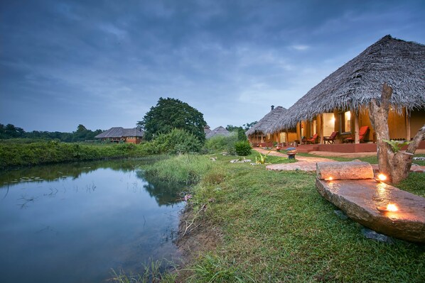 A thatched-roof bungalow with a pond in the foreground, surrounded by lush greenery. - Amuna Ayurveda and Wellness Retreat (Sigiriya)