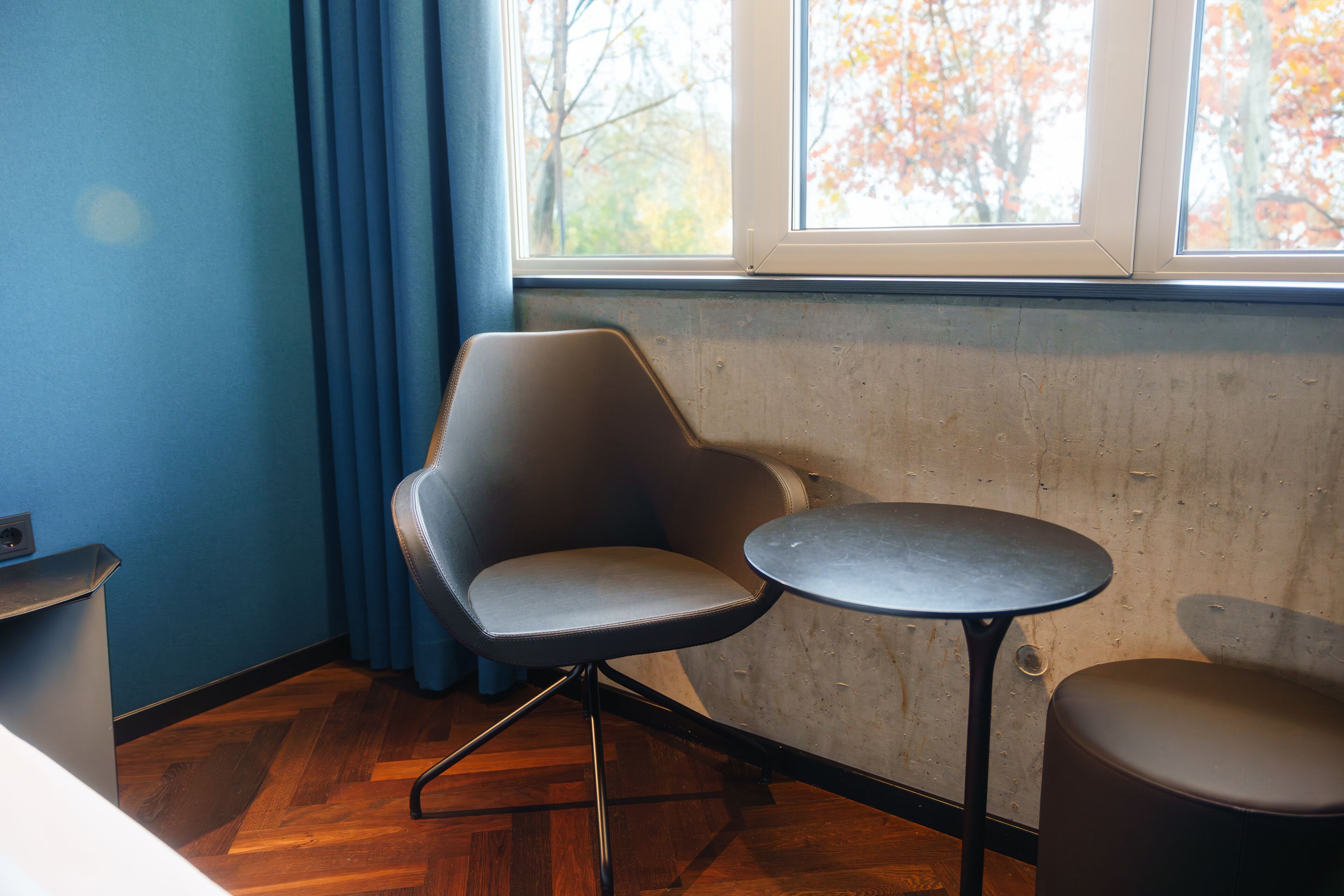 A corner room with a blue wall, a wooden floor, a grey chair, a small round table, and a window with autumn leaves outside.