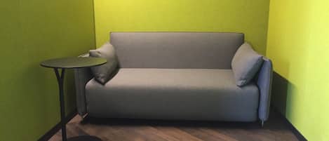Una habitación pequeña y cerrada con un sofá gris y una mesita redonda. Las paredes están pintadas de verde y el suelo tiene un patrón de espiga.