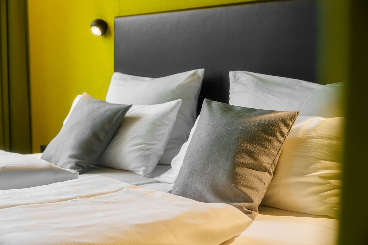 A neatly made bed with white linens and grey pillows, a black headboard, and a yellow wall.