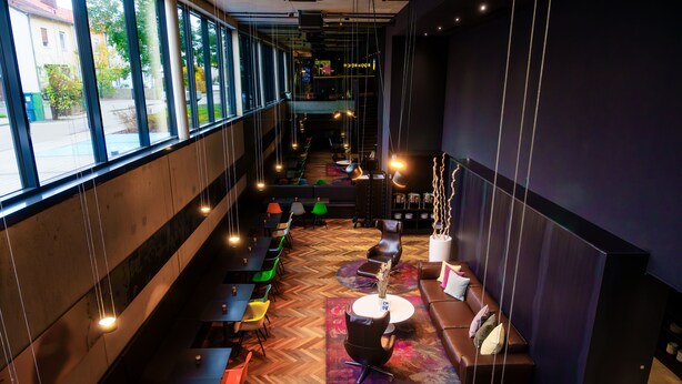 A modern interior with a herringbone wood floor, a central seating area with a round table, and a row of tables and chairs along the walls. - Roomreich (Ingolstadt)