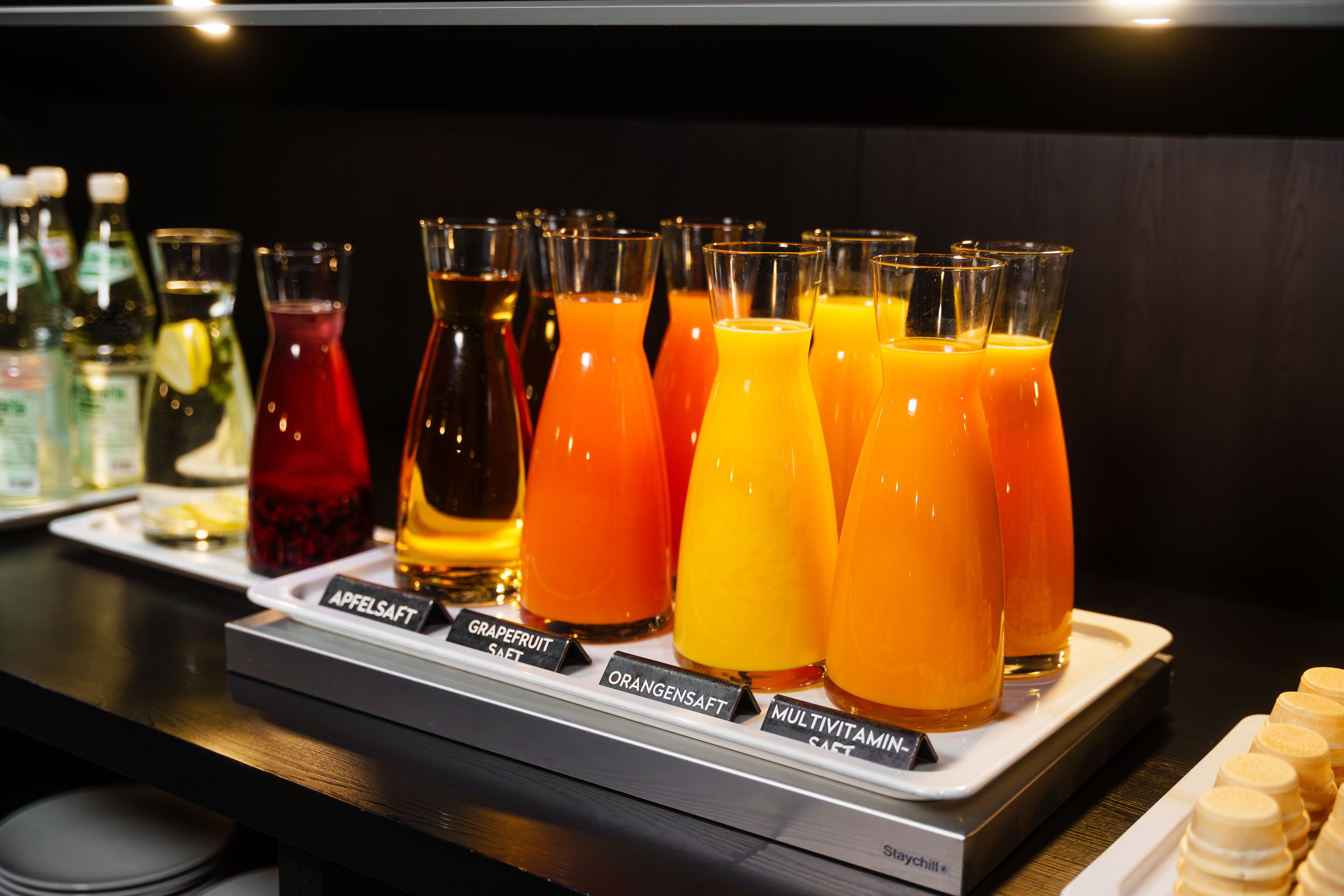 Zona de bar con varios dispensadores de zumo etiquetados con diferentes sabores como pomelo, naranja y multivitamínico.