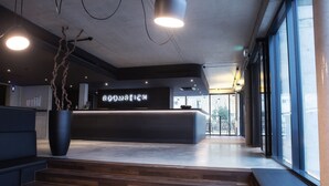 A modern reception area with a dark reception desk, a large potted plant, and a staircase leading to upper floors. - Roomreich (Ingolstadt)