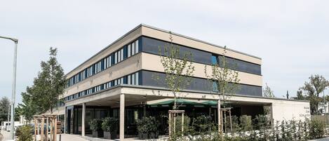 A modern building with a flat roof, large windows, and a covered entrance area with potted plants.