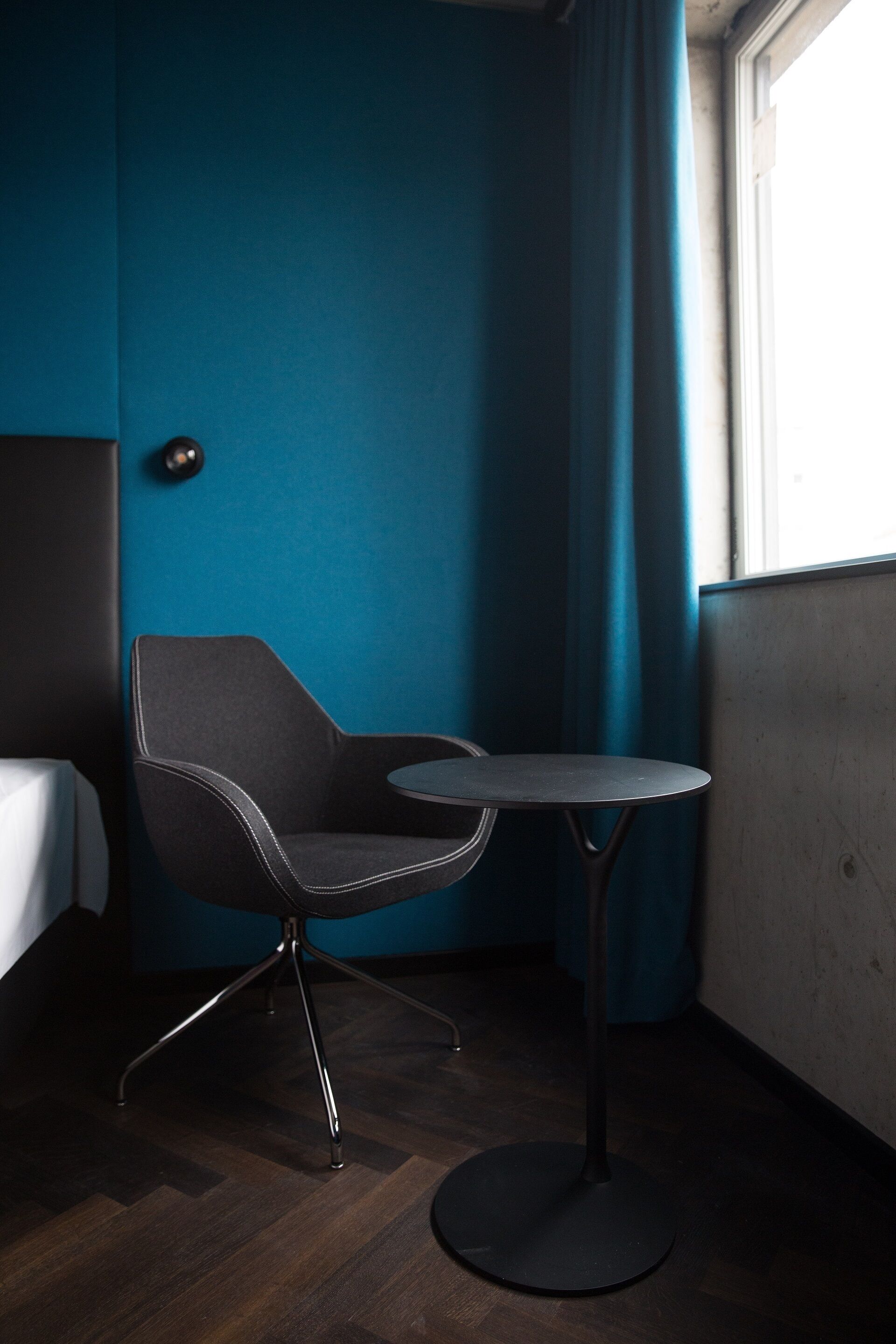 Habitación de esquina con pared azul oscuro, una silla con patas metálicas, una mesita redonda y una ventana con cortinas.