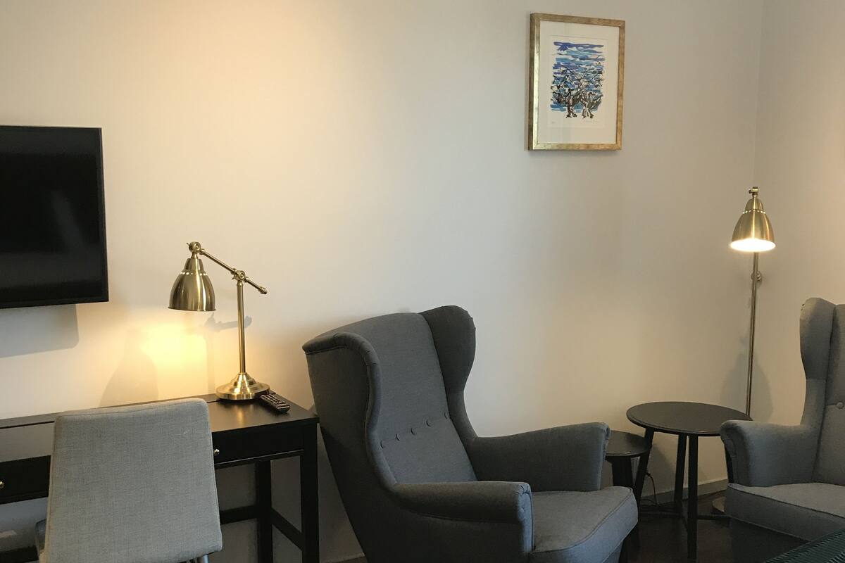 Habitación de hotel con cama, dos sillones, un escritorio con lámpara, televisor y un cuadro en la pared.