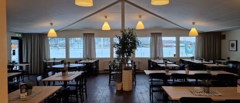 A restaurant with multiple tables and chairs, large windows, and hanging pendant lights.