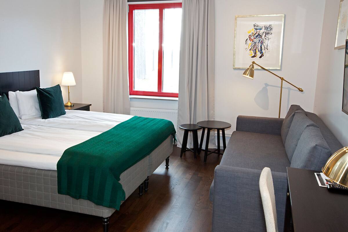 Una habitación de hotel con una cama, un sofá, una mesita y una ventana con marcos rojos.