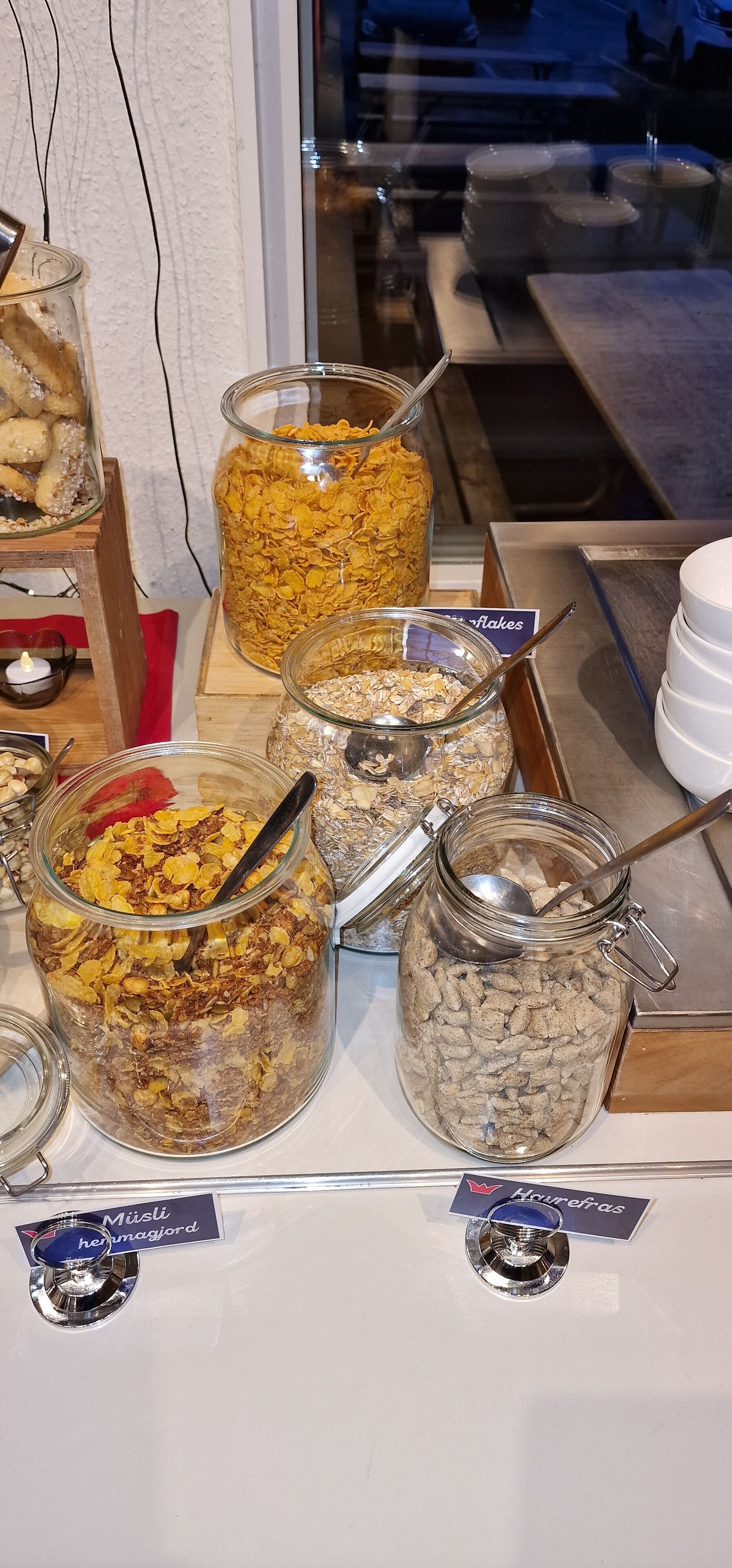 A breakfast buffet with various cereal options in jars, a candle, and a sign indicating 'Muisli' and 'Horefras'.