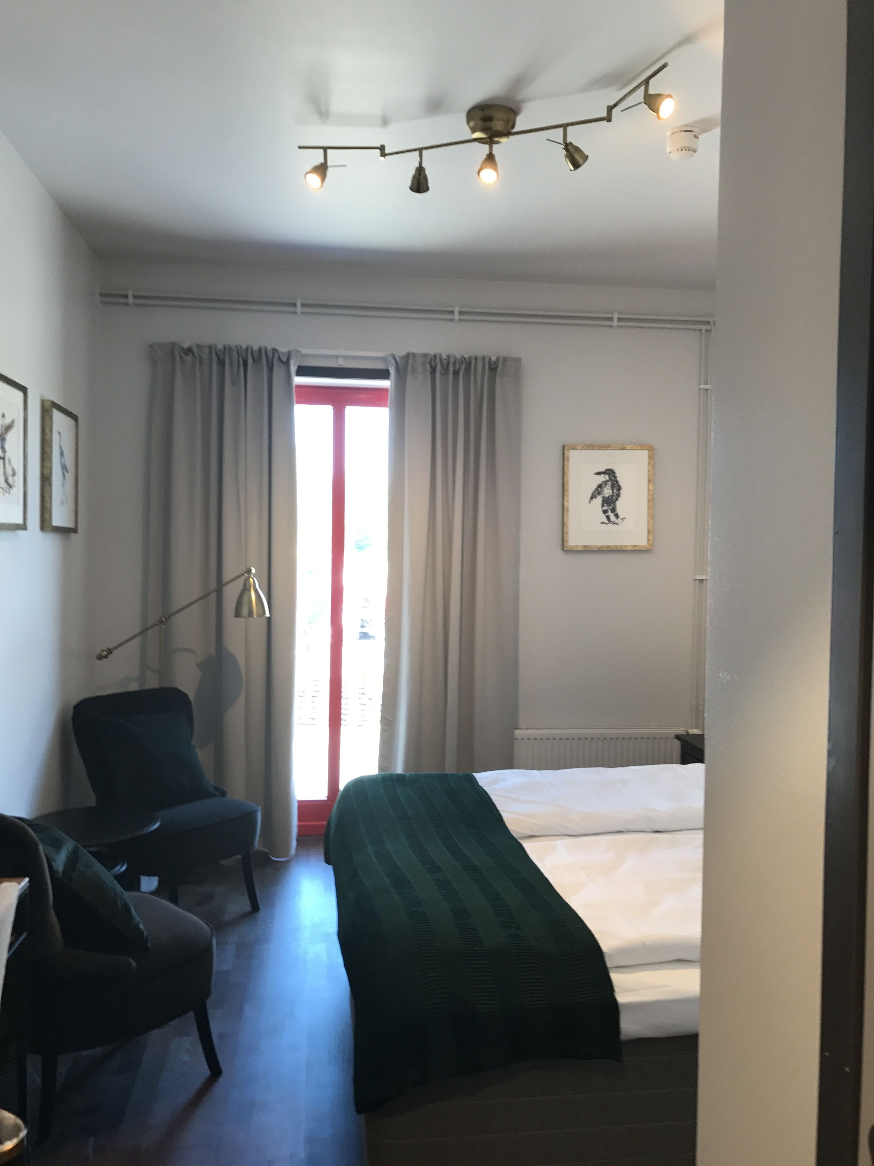 A hotel room with a bed, a chair, a desk, a window with curtains, a ceiling fan, and a framed picture on the wall.
