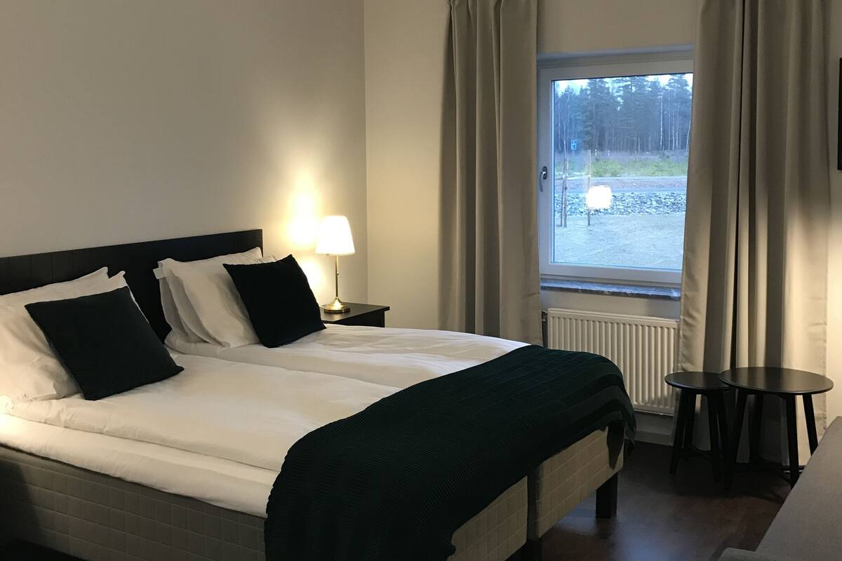 Habitación de hotel con cama, mesitas de noche, lámpara, ventana con cortinas y vistas al exterior.