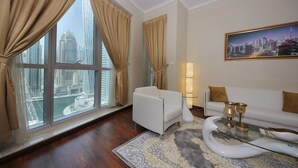 A modern living room with a white sofa, a glass coffee table, and a view of a cityscape through large windows with beige curtains. - Piks Key - Marina Torch Tower (Dubai)