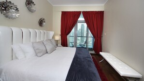 A hotel room with a bed, a bench, and a view of a cityscape through large windows with red curtains. - Piks Key - Marina Torch Tower (Dubai)
