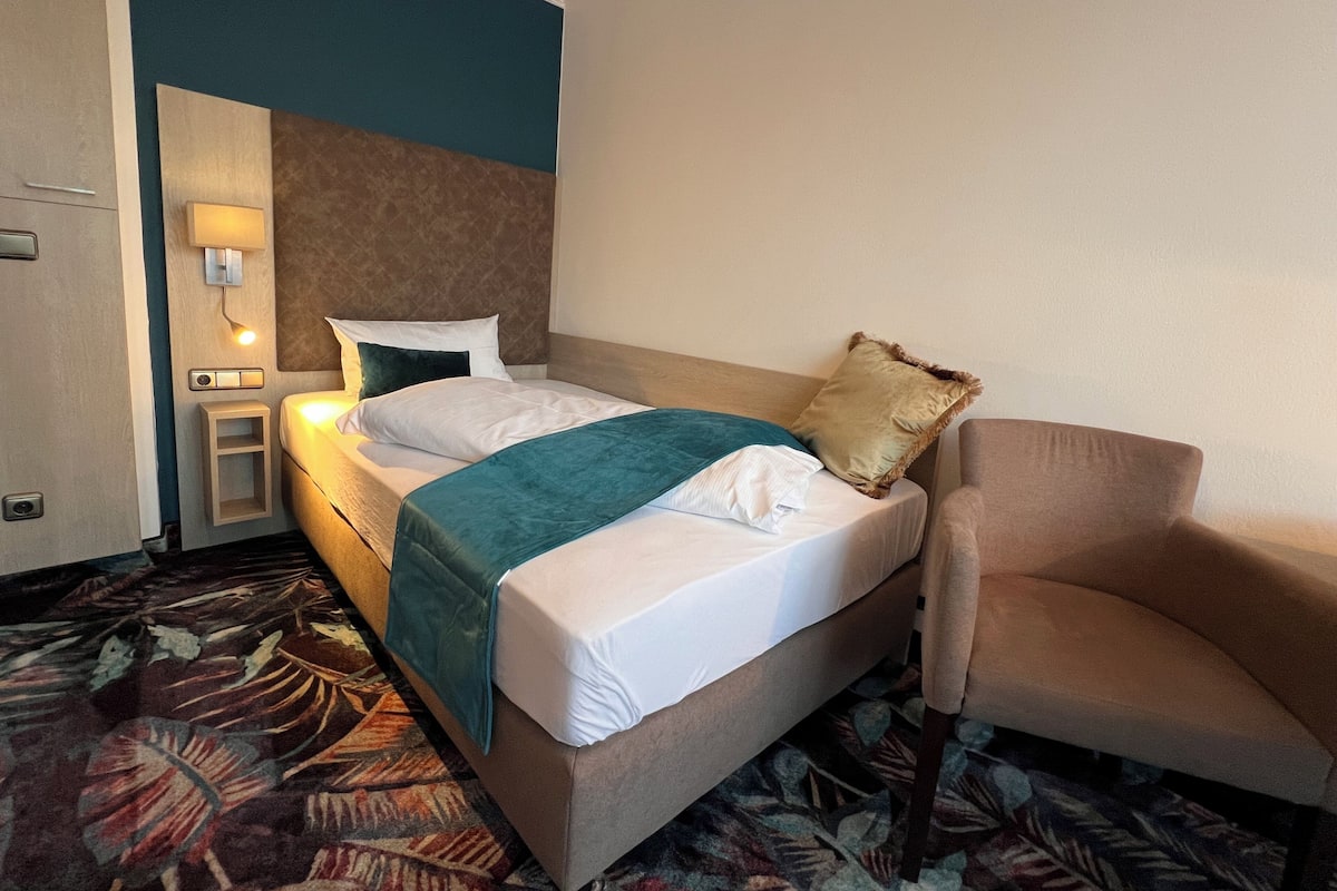 A hotel room with a bed, a chair, a nightstand, and a wall-mounted lamp.