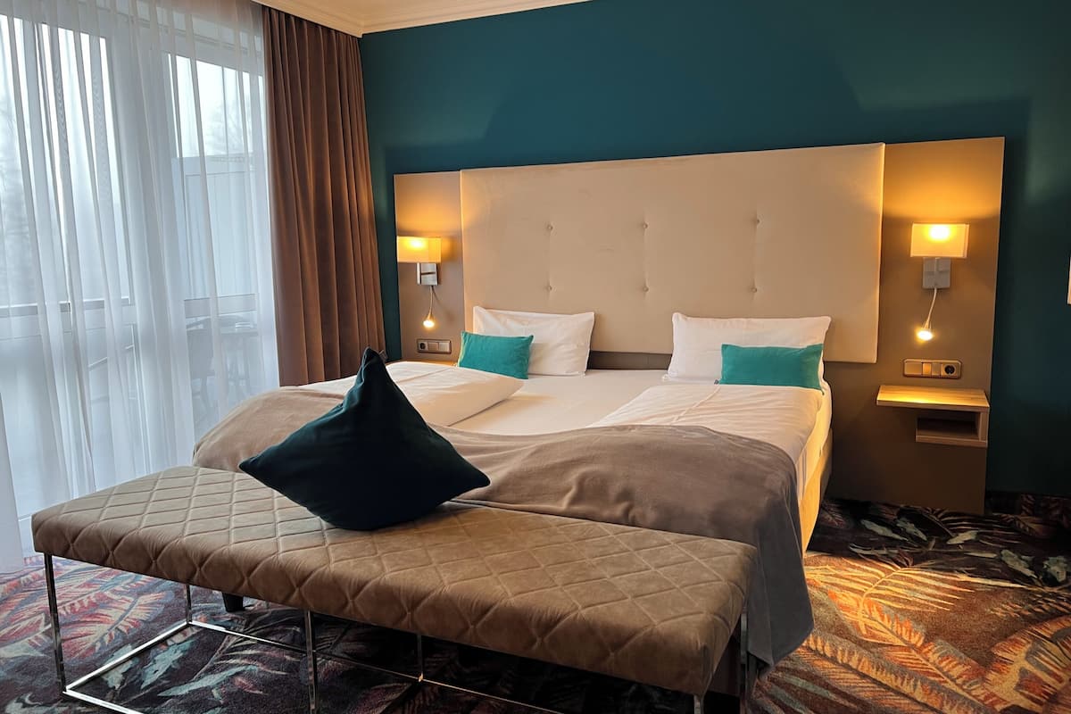 A hotel room with a bed, two bedside tables with lamps, a bench, and a view of the outdoors through sheer curtains.