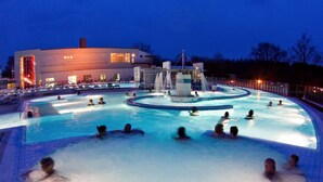 Ampia piscina all'aperto con diverse corsie e una fontana al centro, circondata da lettini e un edificio con finestre illuminate.