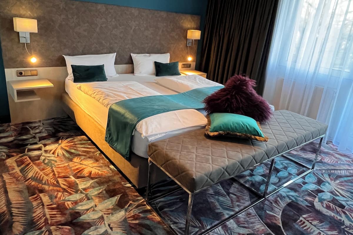 A hotel room with a large bed, two bedside tables, a headboard, and a window with curtains.