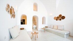 A modern, minimalist living room with a sofa, a small round table with cups, and a large arched doorway leading to a bedroom. - Iriana Suites (Santorini)