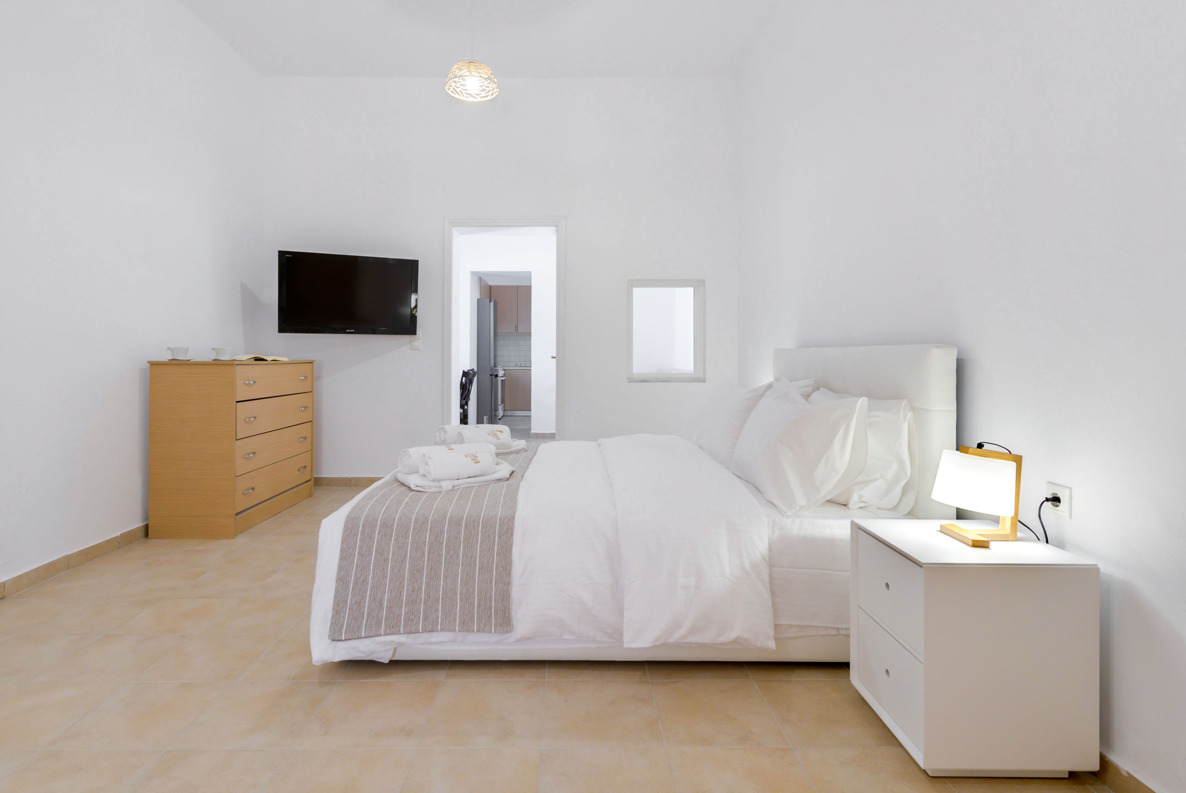 Un dormitorio con una cama, una mesita de noche con lámpara, un aparador de madera y un televisor montado en la pared.