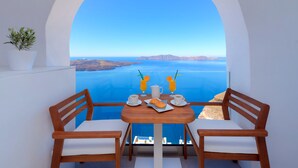 A wooden table with two chairs, a potted plant, and a view of the sea through an arched window. - Iriana Suites (Santorini)