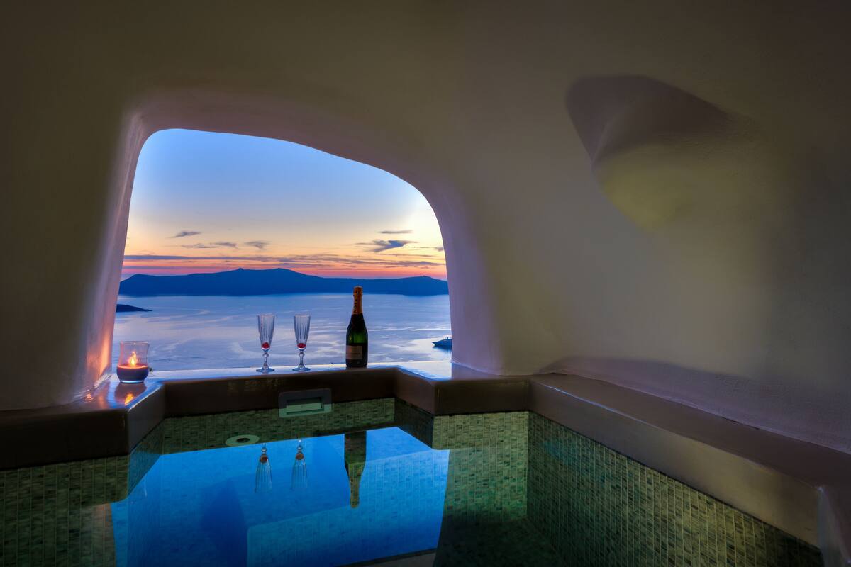 Vista desde una habitación con ventanal de vidrio que da a un cuerpo de agua y montañas al atardecer.