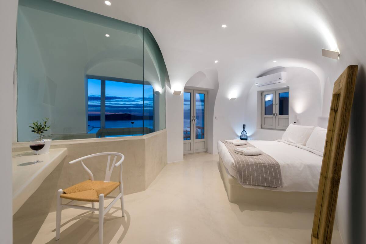 Un dormitorio moderno con un ventanal grande, una cama, una silla y una mesita con una planta y una copa de vino.