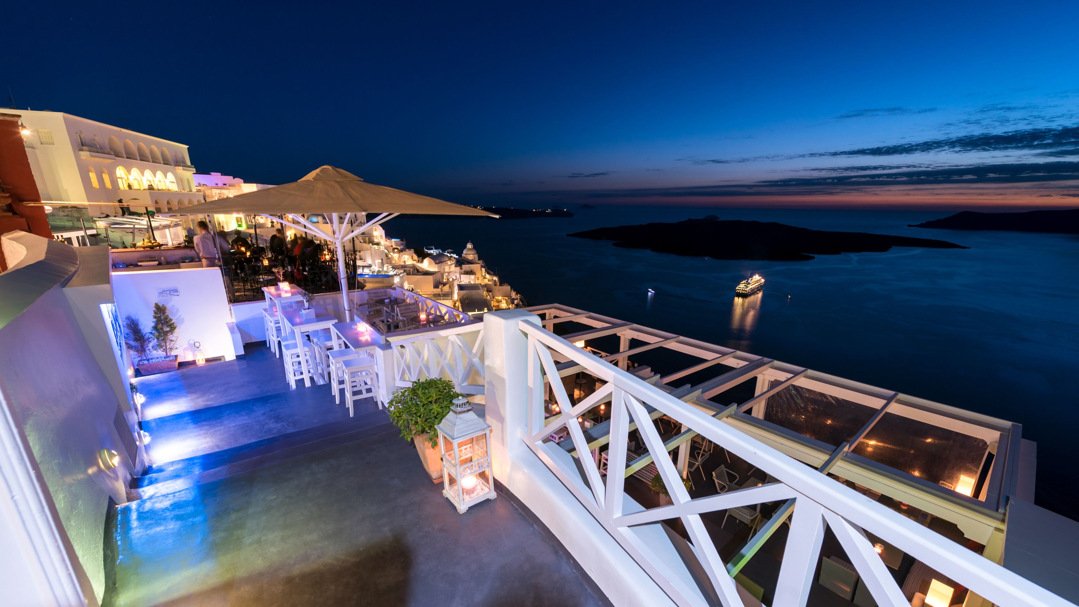A rooftop bar with white furniture, a view of the sea, and a building with arches in the background.