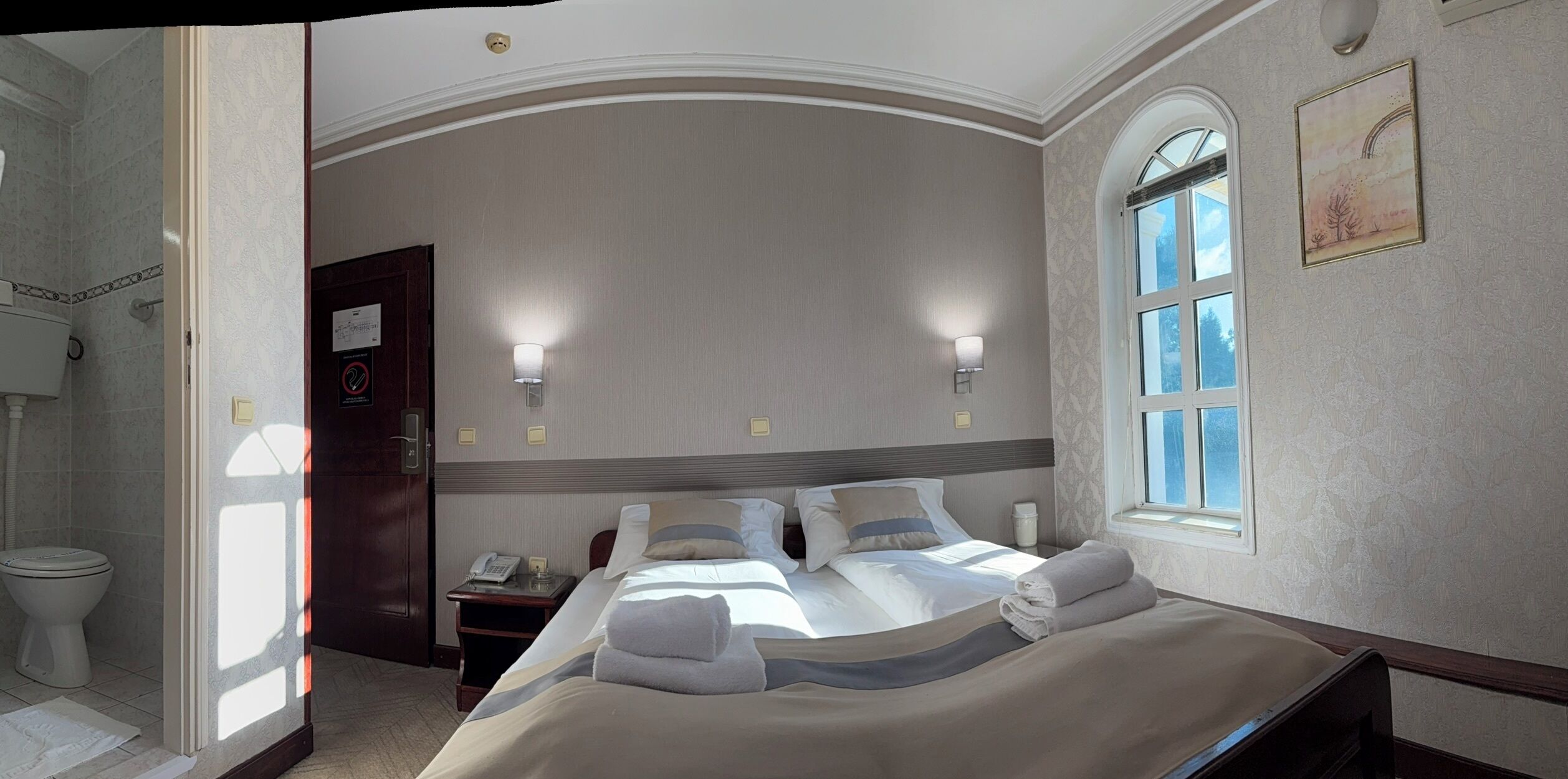 A hotel room with a large bed, two bedside tables, a nightstand, a window with a view, and a door leading to a bathroom.