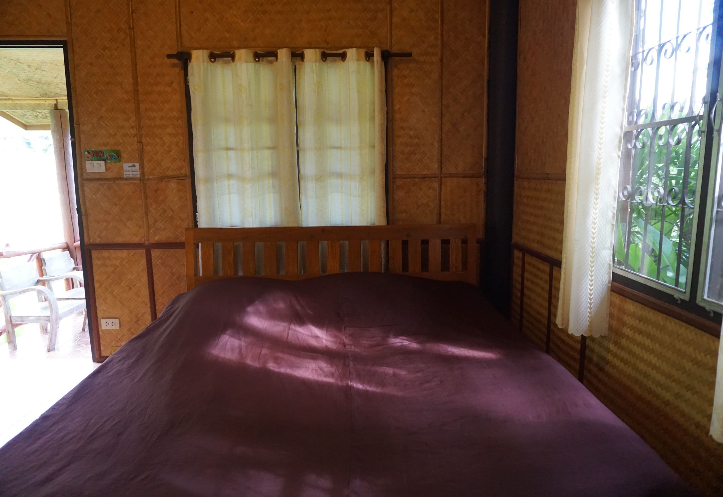 A room with a wooden bed, a window with metal bars, and a view of greenery outside.