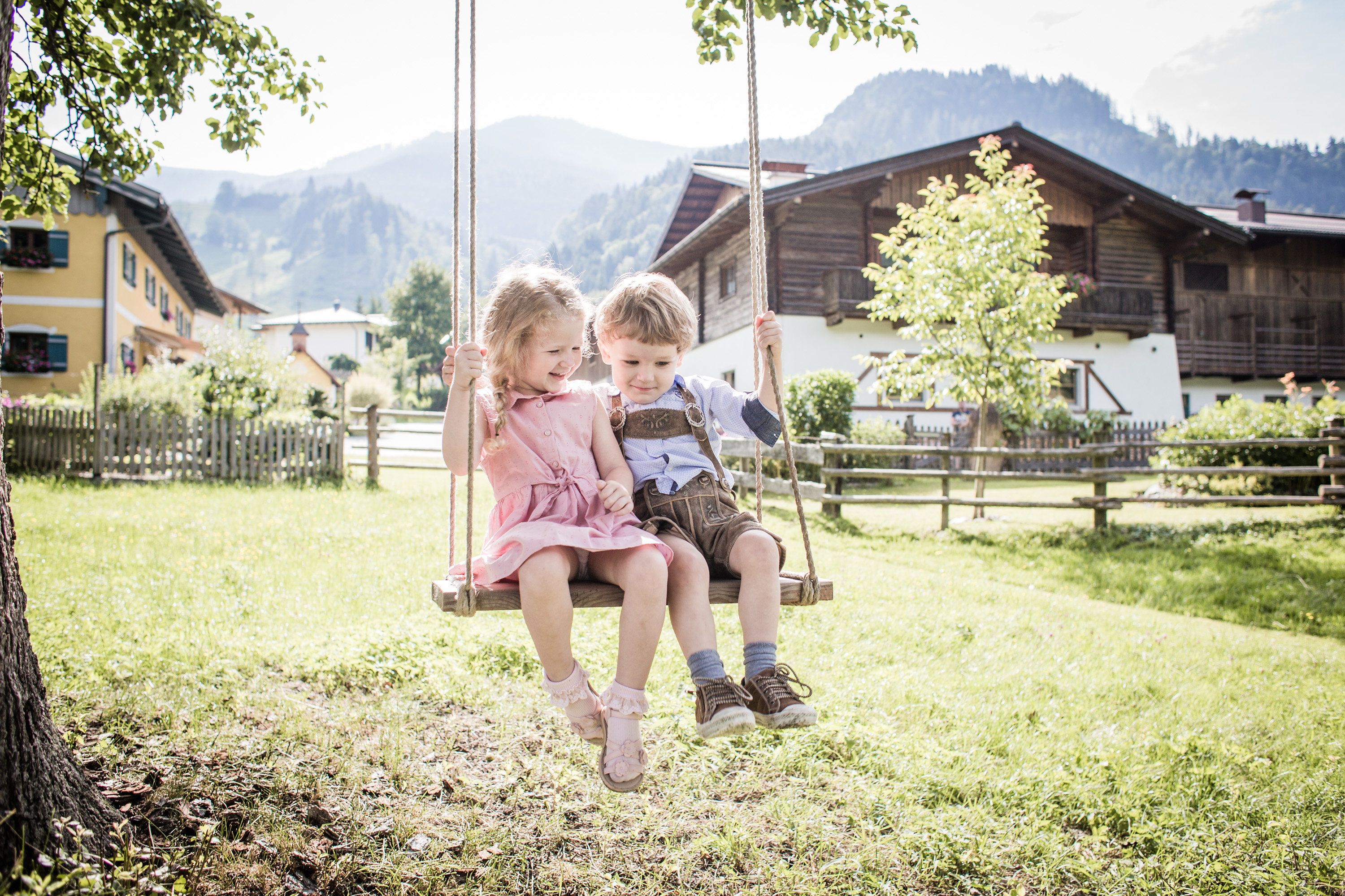 Two children on a swing in a rural setting with wooden houses and mountains in the background.