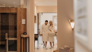 A couple in white robes in a modern bathroom with a wooden accent wall, a glass shower enclosure, and a small table with towels.