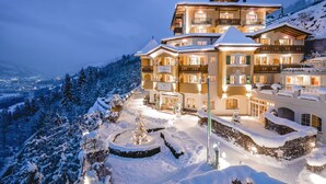 A multi-story hotel with balconies, a snow-covered garden, and a view of a snowy mountain range.