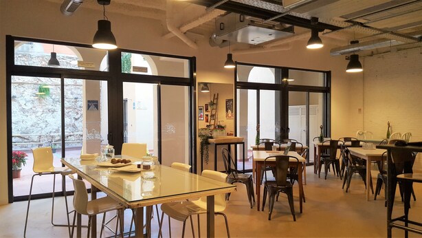 A modern dining area with glass tables and chairs, large windows, and hanging pendant lights. - Ten To Go Hostel (Barcelona)