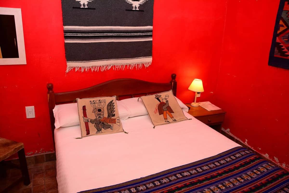 Uma cama com duas almofadas, uma mesa de cabeceira com abajur, e uma parede vermelha com uma tapeçaria pendurada.