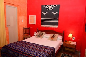 A room with a red wall, a bed with a patterned bedspread, a wooden nightstand with a lamp, and a wall hanging with a bird design. - Hostal Yatasto (Salta)