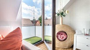 A modern room with a large sliding glass door leading to a balcony, a wooden circular bench, and a white bedside table with a glass container. - Appartmenthaus am Erlbach (Buch am Erlbach)