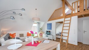 A modern loft-style living space with a wooden loft bed, a dining area with a wooden table, and a cozy seating area with a sofa and cushions. - Appartmenthaus am Erlbach (Buch am Erlbach)